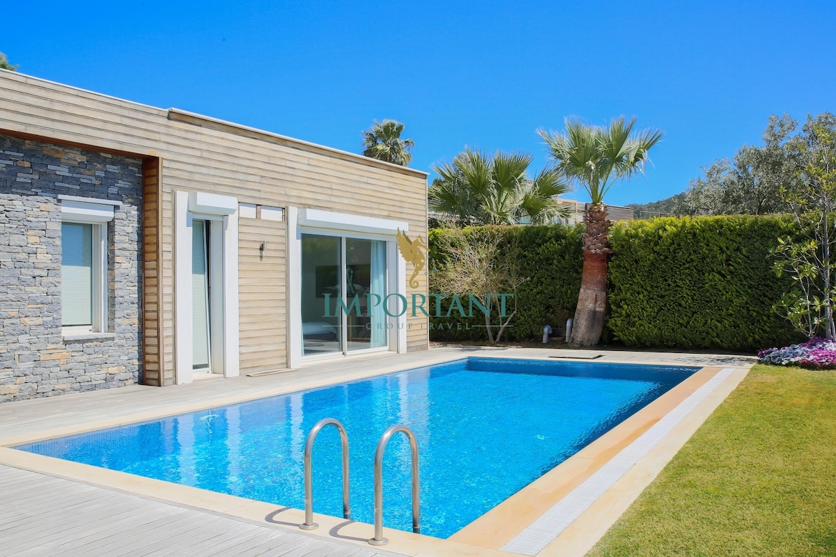 A private pool is framed by a wooden deck, surrounded by lush greenery and palm trees. Large glass doors from the villa offer an inviting view of the pool area, providing easy access to the outdoor space. A vibrant garden adds to the tranquil setting.