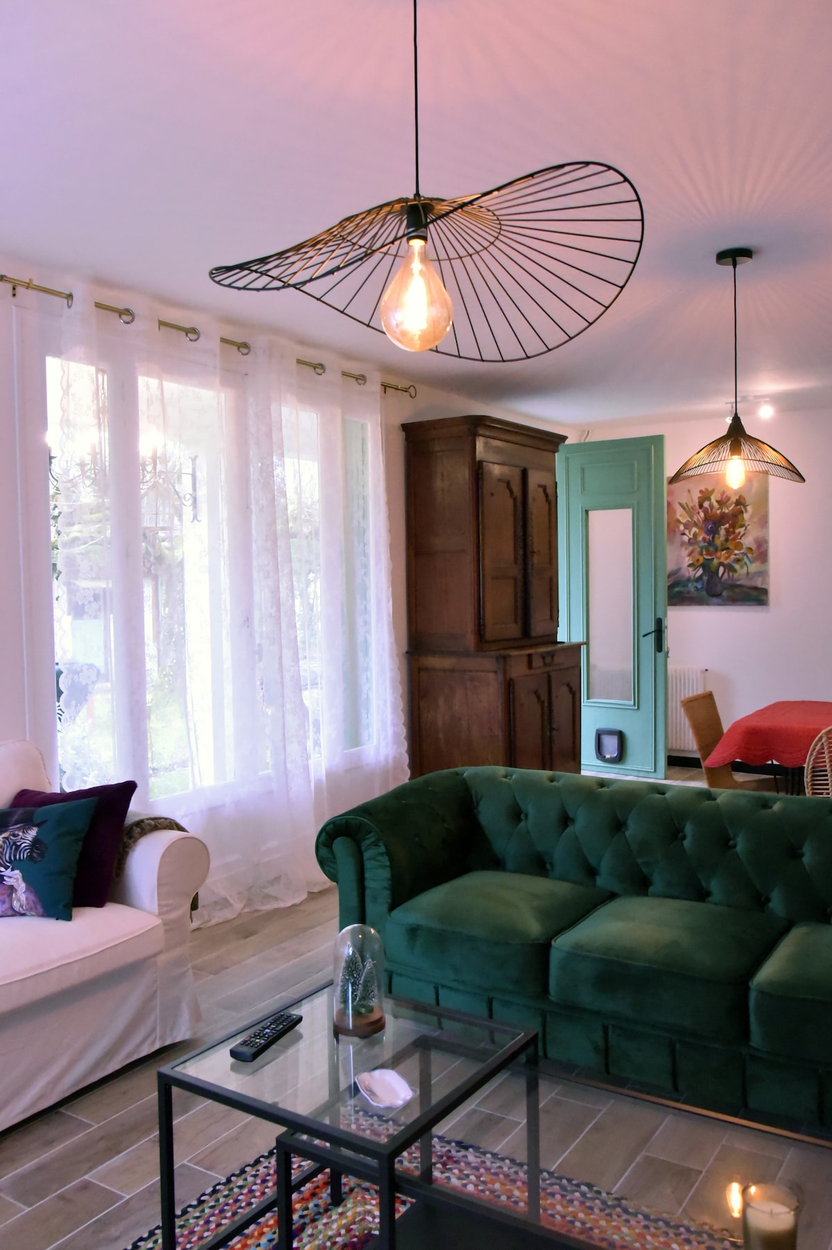 A bright living area features large windows dressed in sheer white curtains, allowing natural light to fill the space. A green tufted sofa complements a contrasting white armchair. A vintage wooden cabinet and contemporary light fixtures add character to the room, enhancing its cozy atmosphere.