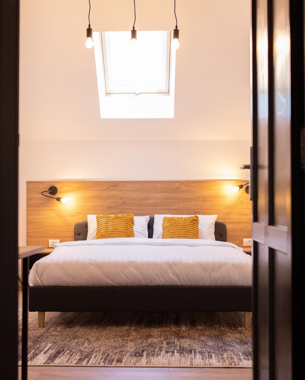 A well-lit bedroom features a spacious bed with soft linens and decorative cushions. Warm light fixtures are mounted on the wall beside the bed, with a skylight above allowing natural light to brighten the space. A stylish area rug covers the floor.