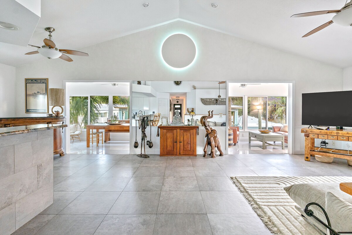 The spacious open-concept living area features high ceilings with a circular light fixture and is adorned with minimalist decor. Large windows provide ample natural light and offer views of the outdoor space. A comfortable seating area and wooden dining table are visible.