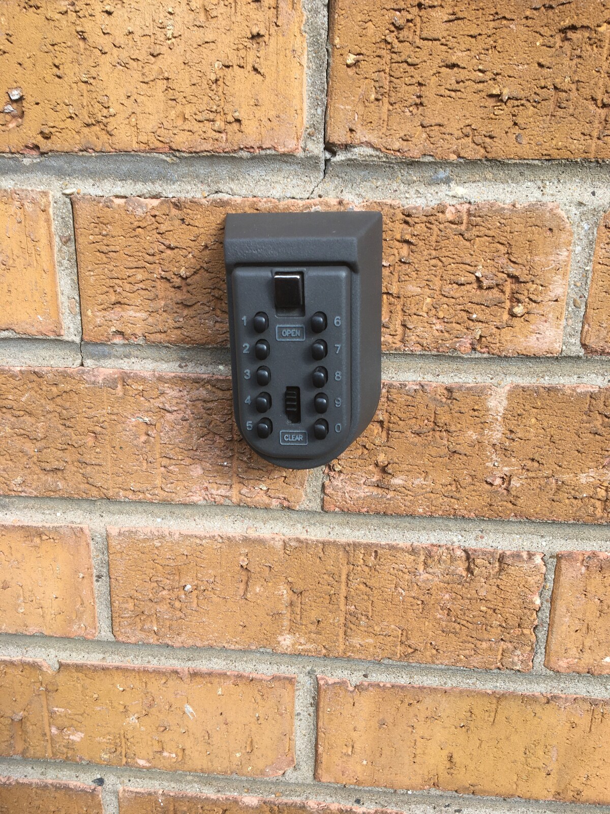 A secure keyless entry lockbox is mounted on a brick wall, featuring a numerical keypad for code entry. The surrounding bricks are a muted orange color, providing a textured backdrop that emphasizes the lockbox.