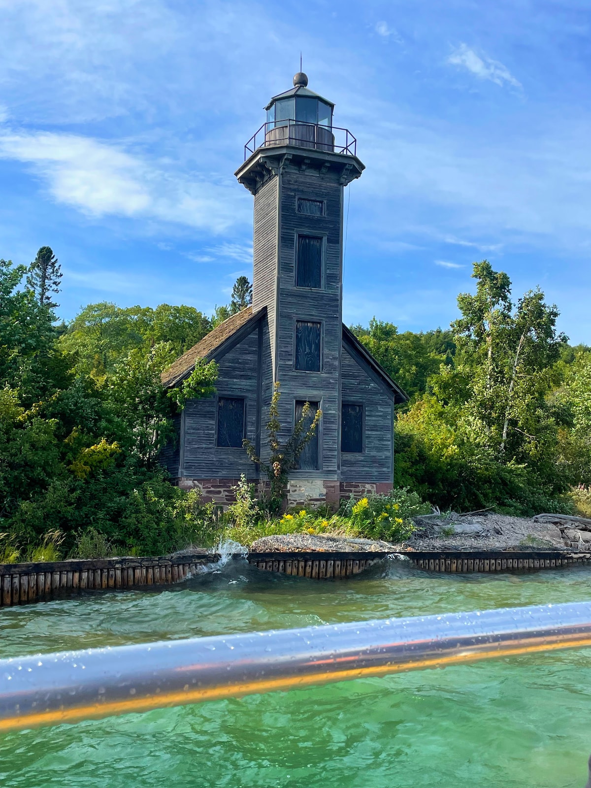 A tall, vintage lighthouse rises amidst lush greenery, surrounded by trees and shrubs. Its weathered gray exterior contrasts with the bright blue sky. The calm water gently laps at the rocky shoreline, adding a serene quality to the scene.