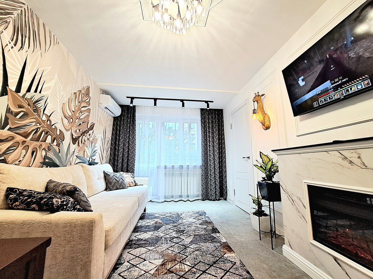 A living area features a comfortable light-colored sofa with decorative cushions, complemented by a large area rug. Natural light enters through the windows dressed in sheer curtains. A flat-screen Smart TV is mounted on the wall, above a modern electric fireplace.