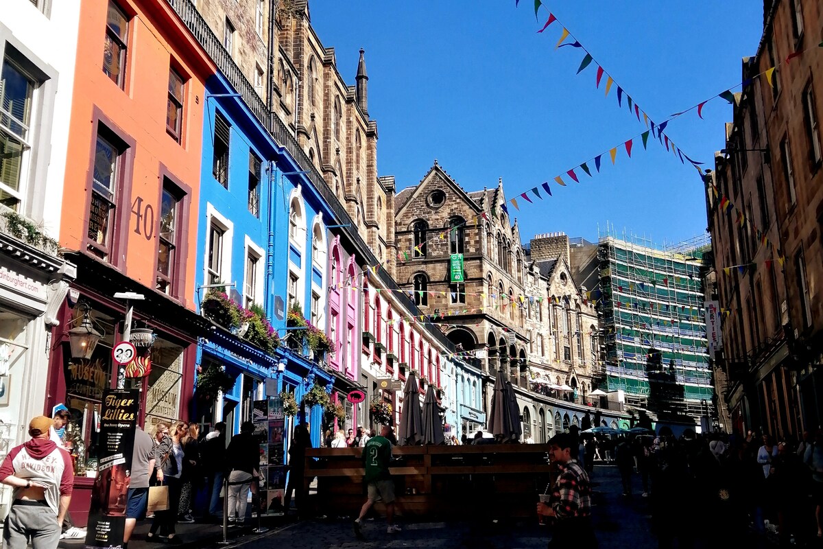 A vibrant street scene is captured, showcasing colorful buildings painted in shades of orange, blue, and pink. Banners are strung overhead, creating a festive atmosphere. People stroll along the cobblestone path, with scaffolding visible on one building in the background.