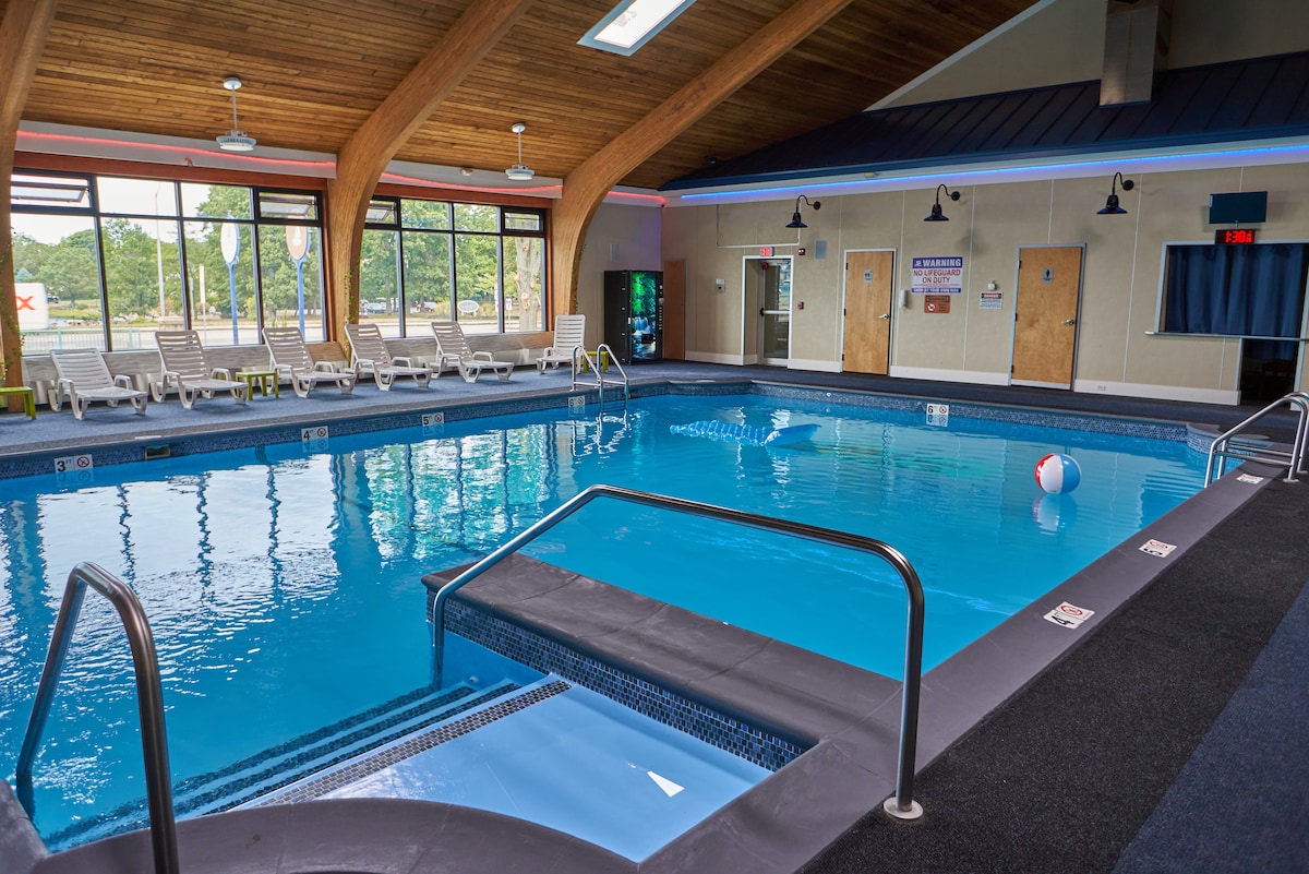 An indoor swimming pool area features a clear blue water expanse, surrounded by lounge chairs. Large windows offer natural light, while an inviting ambiance is created by wooden beams overhead. A poolside ramp provides accessibility, and a beach ball floats gently on the water's surface.