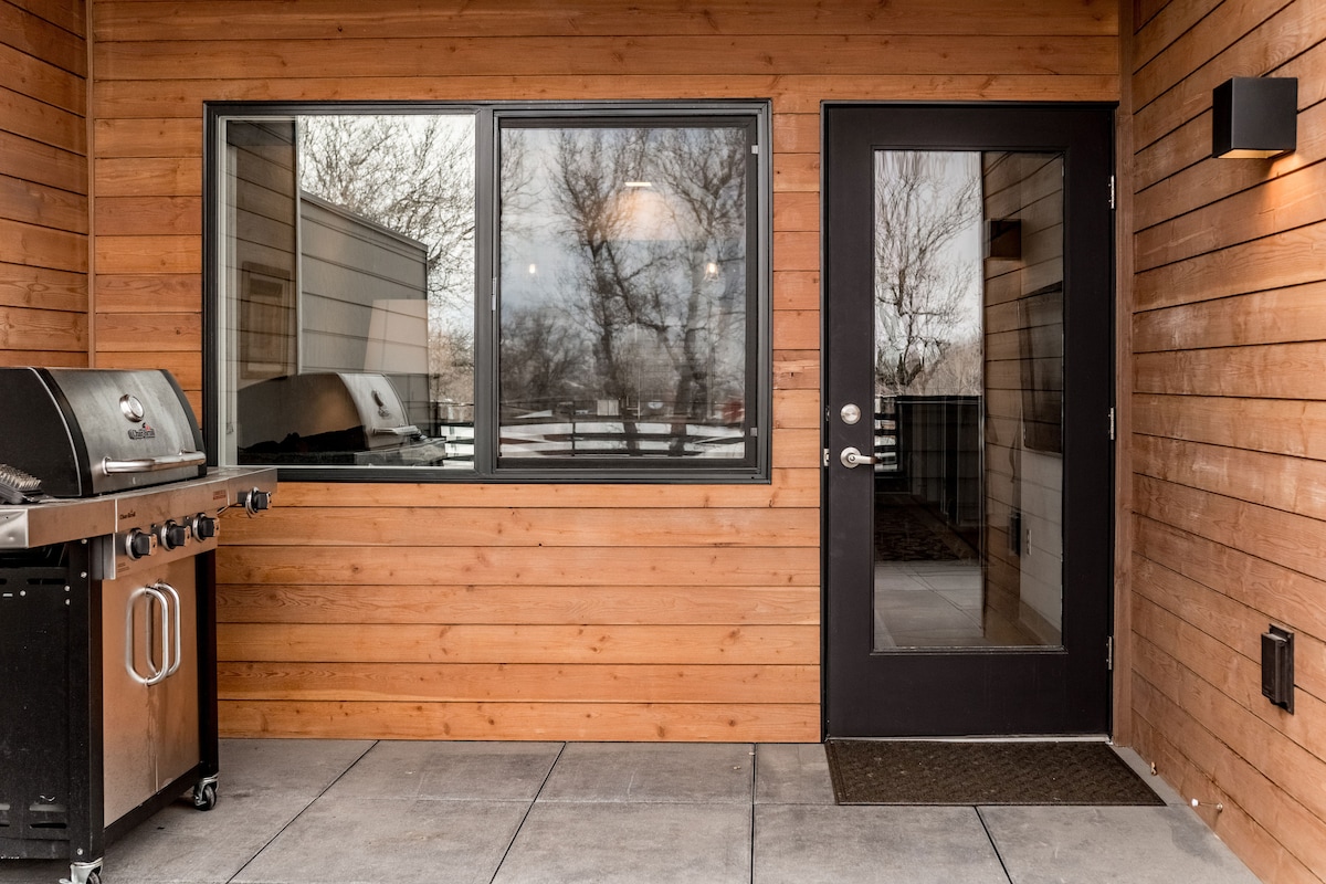 A wooden exterior wall highlights the entrance to the condo, featuring a dark door. A large window reflects the outdoor scenery. An outdoor grill is positioned adjacent to the door, providing convenient access for cooking.