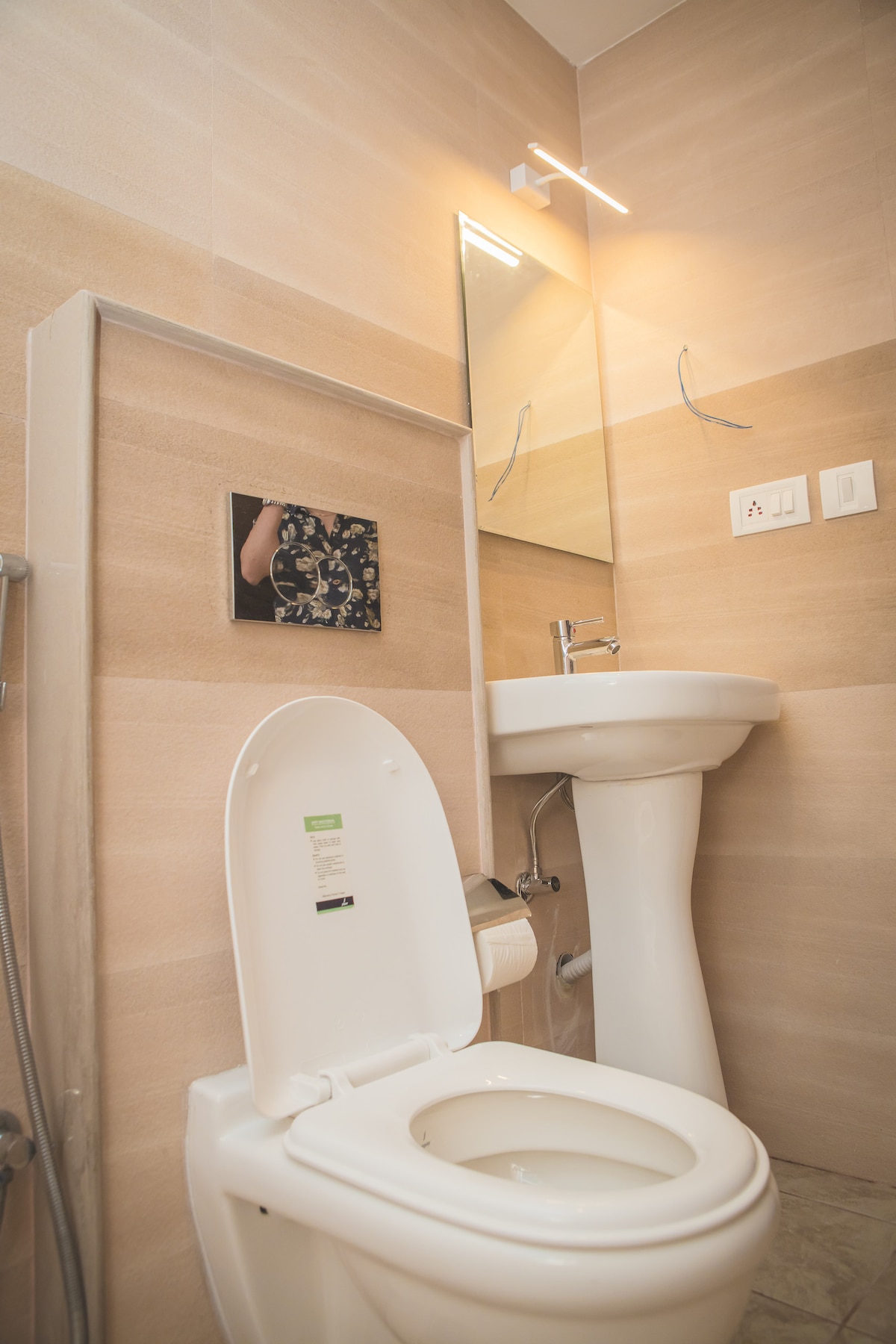 A modern bathroom features neutral-toned tiles and a compact design. A wall-mounted mirror is positioned above a sleek pedestal sink, and a contemporary toilet occupies the space. Soft lighting highlights the clean lines of the fixtures.