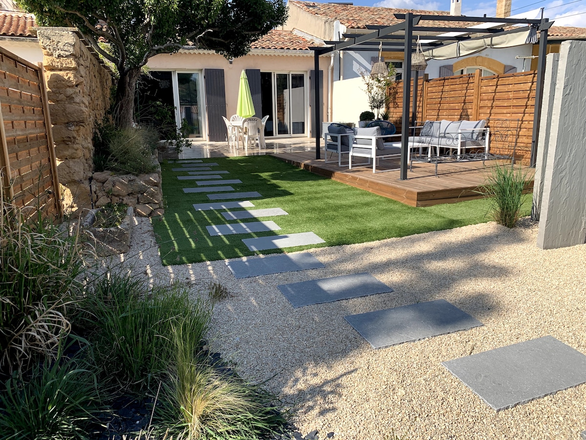 A landscaped garden features a combination of grass and gravel pathways leading to a wooden deck area. Comfortable seating with a table is arranged under a pergola, while a bright green umbrella provides shade. Lush plants and decorative stones enhance the outdoor space.