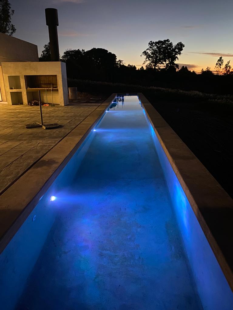 A long, narrow pool stretches under a dusky sky, illuminated by soft blue lights. The stone surrounding is smooth and reflects the twilight colors, while a built-in grilling area is visible nearby, enhancing the outdoor space for gatherings.