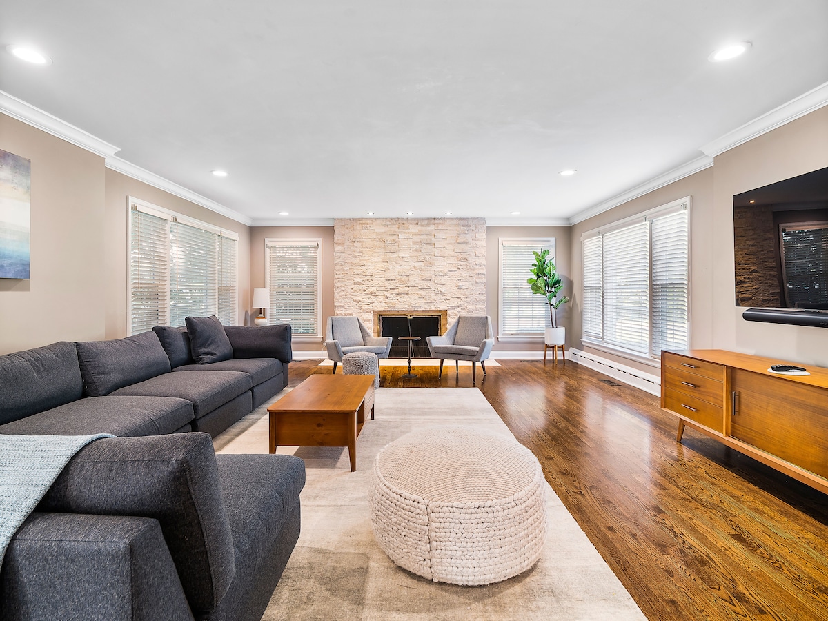 A spacious living area features a large sectional sofa and matching pouf, complemented by a wooden coffee table. A stone fireplace serves as a focal point, while two armchairs and a sleek television provide additional seating and entertainment options. Natural light filters through multiple windows.