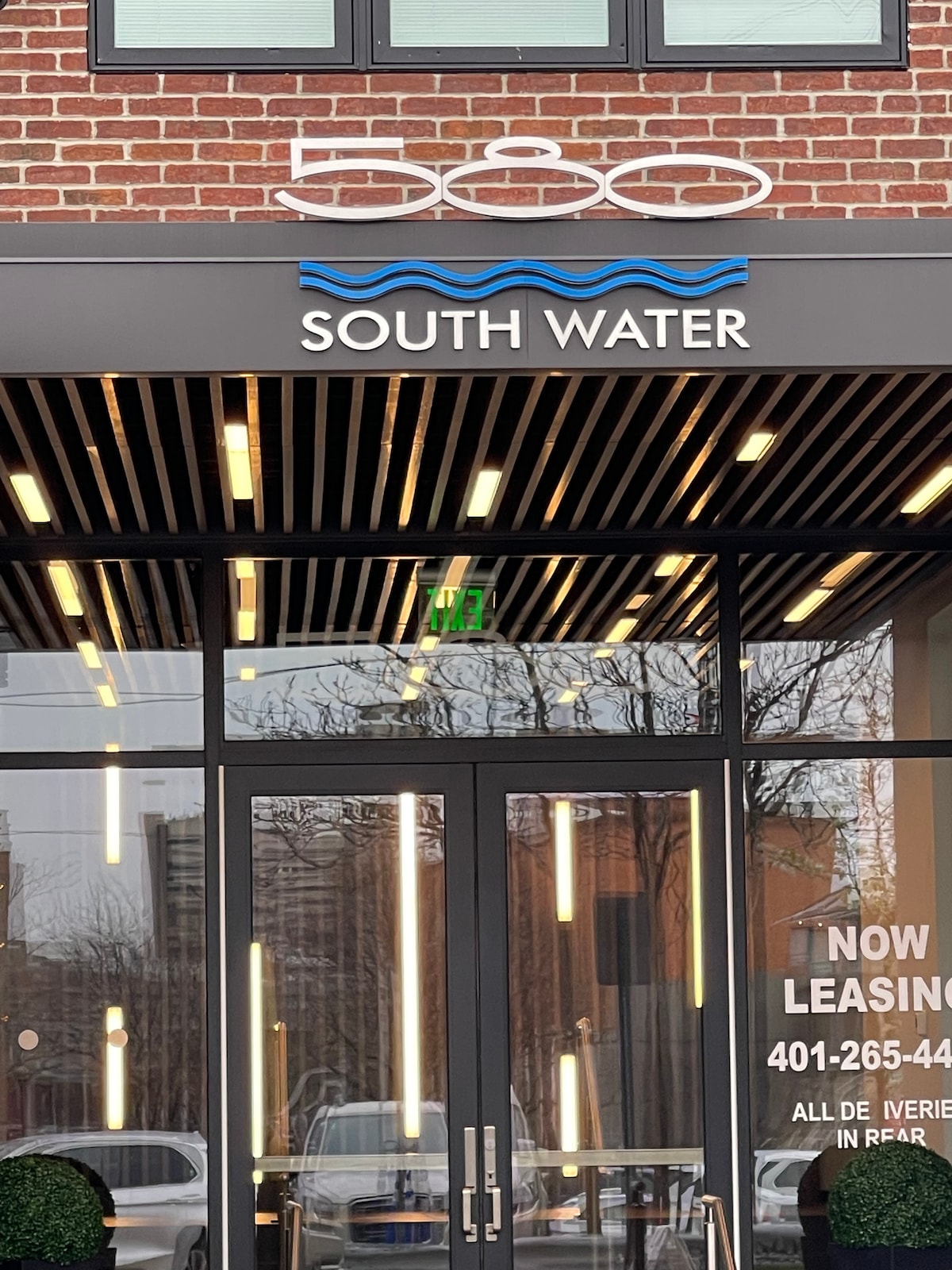 The entrance of a contemporary building is displayed, featuring sleek glass doors and a modern awning. The building name, '580 SOUTH WATER,' is prominently displayed above, accompanied by a wavy graphic. Bright lighting fixtures hang above, creating a welcoming atmosphere.