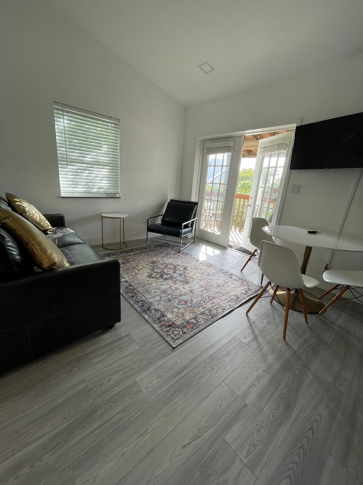 A cozy living area is presented with a modern black sofa adorned with decorative pillows. A glass door leads to a balcony, allowing natural light to fill the space. A small round dining table is positioned next to a chair, complemented by a colorful area rug and a mounted TV.