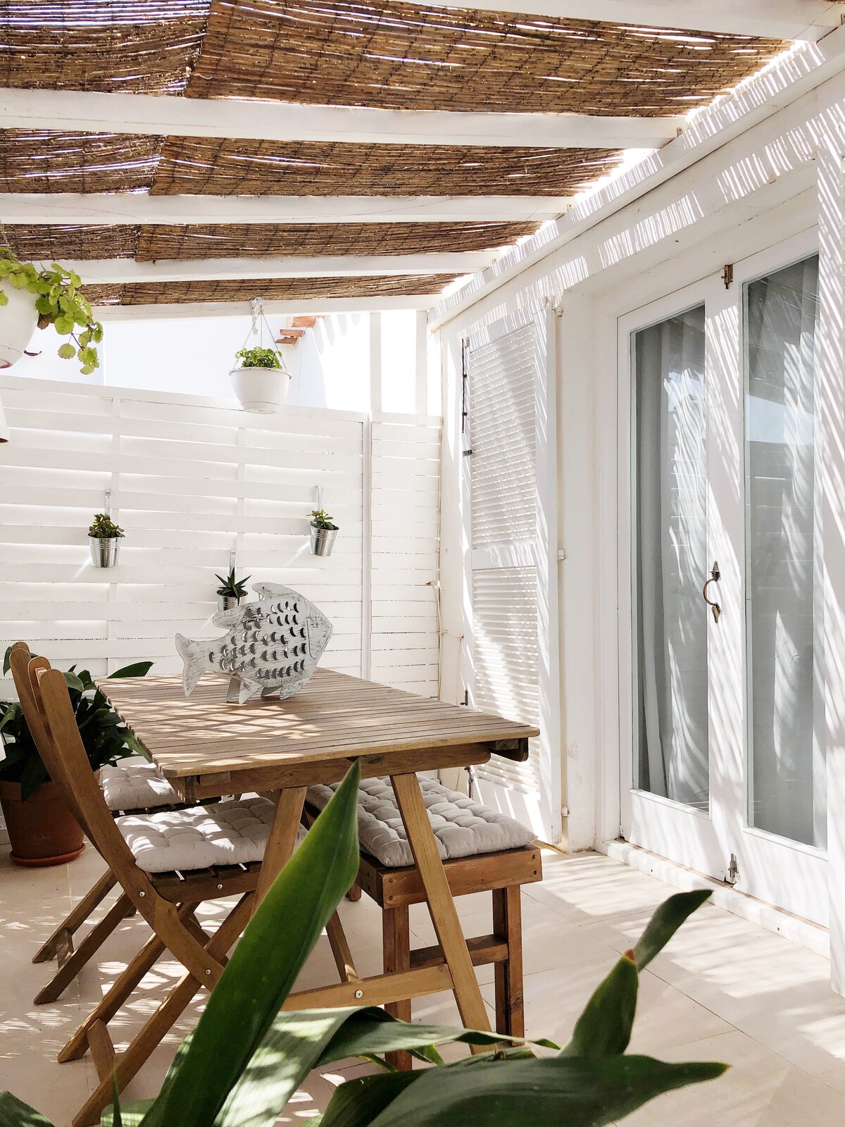 A shaded terrace is presented with a wooden table and folding chairs, surrounded by potted plants. Light filters through a woven canopy, creating a dappled effect on the floor and furnishings. Glass doors lead to the indoor space, enhancing the open atmosphere.