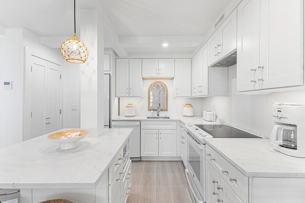 A modern kitchen is presented with white cabinetry and sleek countertops. Bright lighting highlights the room's spacious layout, featuring an island with a decorative bowl. Appliances, including a refrigerator and stove, are integrated into the design, enhancing functionality.