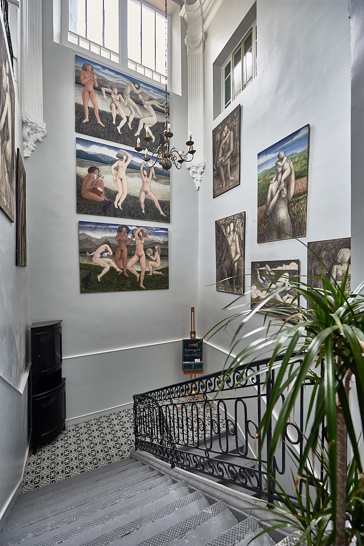 The interior staircase is framed by tall walls adorned with various artworks, featuring human figures and natural landscapes. A chandelier hangs above, providing soft illumination. A decorative plant is positioned beside the staircase, which leads down to a patterned tiled floor.