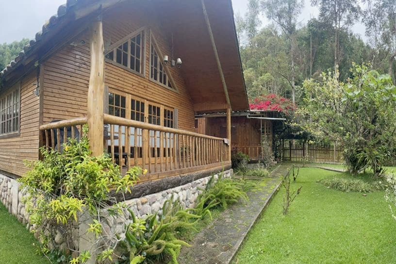 The image depicts a two-story wooden cabin with a large porch, surrounded by lush greenery. A stone pathway leads through the garden, which features various plants and trees. Large windows allow natural light to illuminate the interior, creating a connection with the outdoor environment.