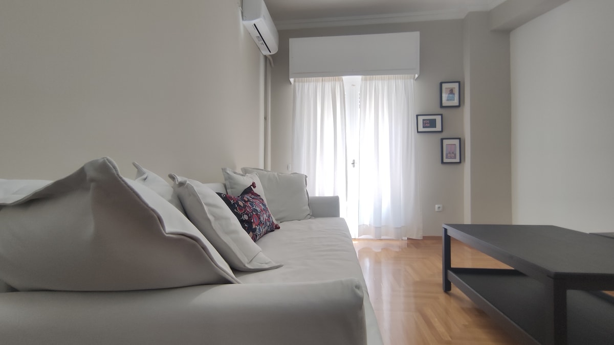 A light-filled living area features a comfortable sofa adorned with decorative cushions. A dark coffee table sits opposite the sofa, while sheer white curtains cloak a glass door that leads to a balcony, providing a bright and airy atmosphere.