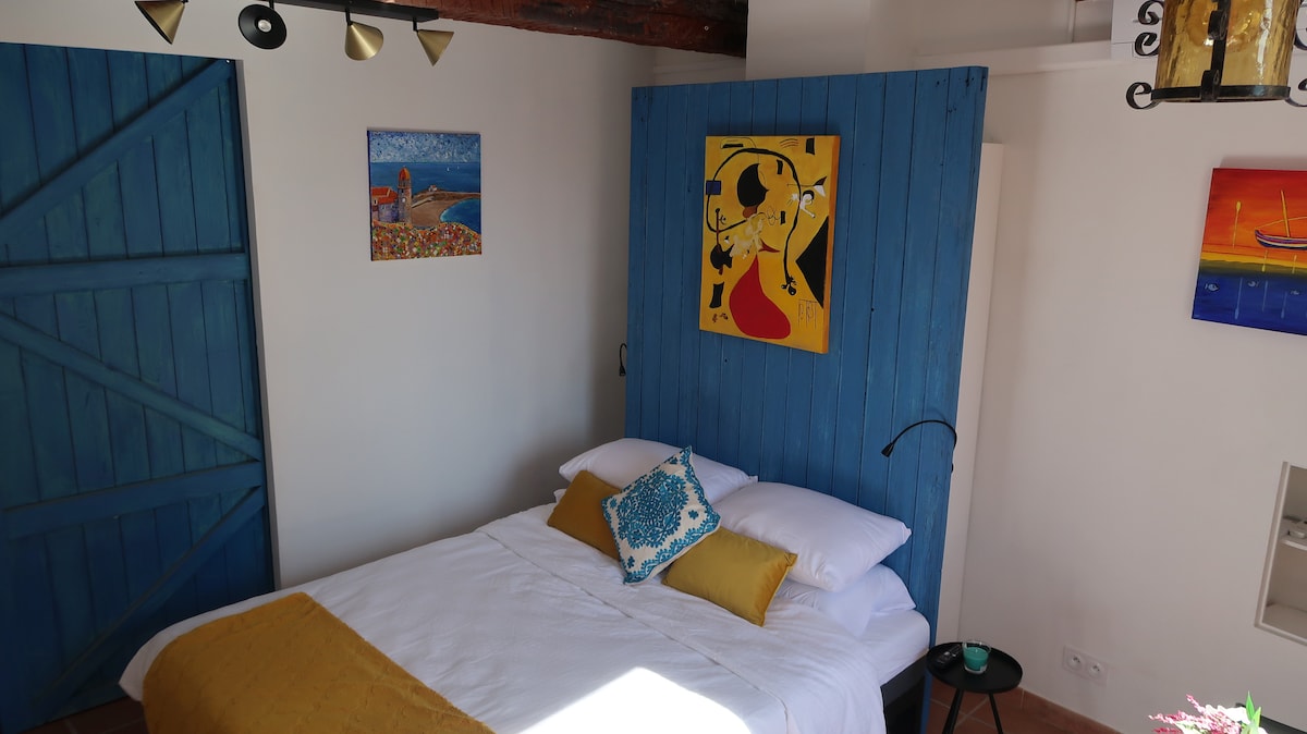 A cozy sleeping area features a queen size bed topped with white linens and vibrant pillows, set against a blue wooden wall. Colorful artwork adorns the walls, enhancing the space's artistic atmosphere, while natural light illuminates the room through a nearby window.