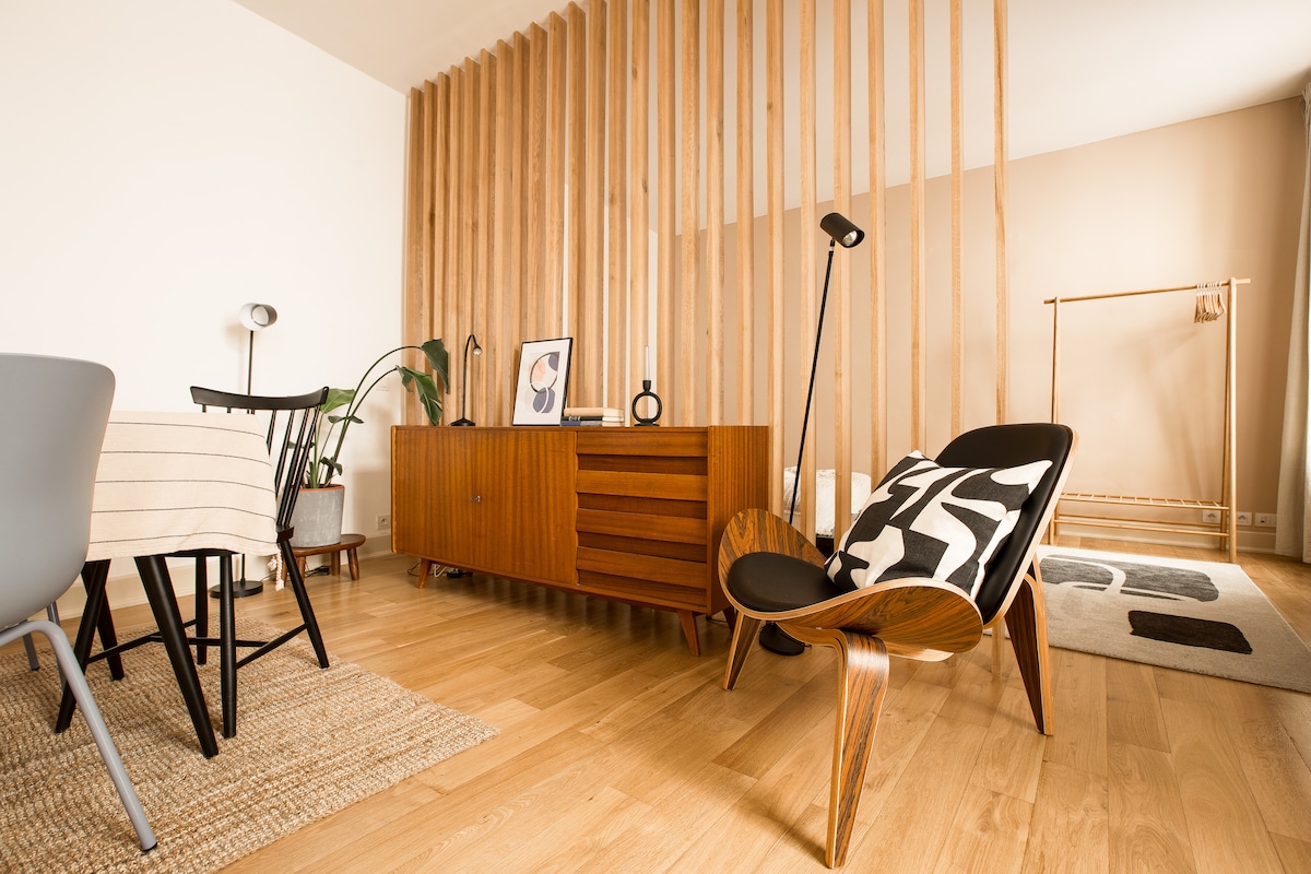 A stylish living area is presented with wooden paneling dividing the space. A light wooden floor complements an eclectic mix of furniture, including a unique chair and a sleek sideboard. Natural light highlights the inviting atmosphere, enhanced by a few decorative elements in the background.