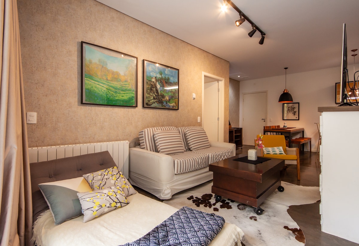 A relaxing living space showcases a combination of a spacious sofa and a comfortable daybed, adorned with patterned cushions. Framed artwork is displayed on the textured walls, while a wooden coffee table rests on a soft area rug, complementing the dining area in the background.
