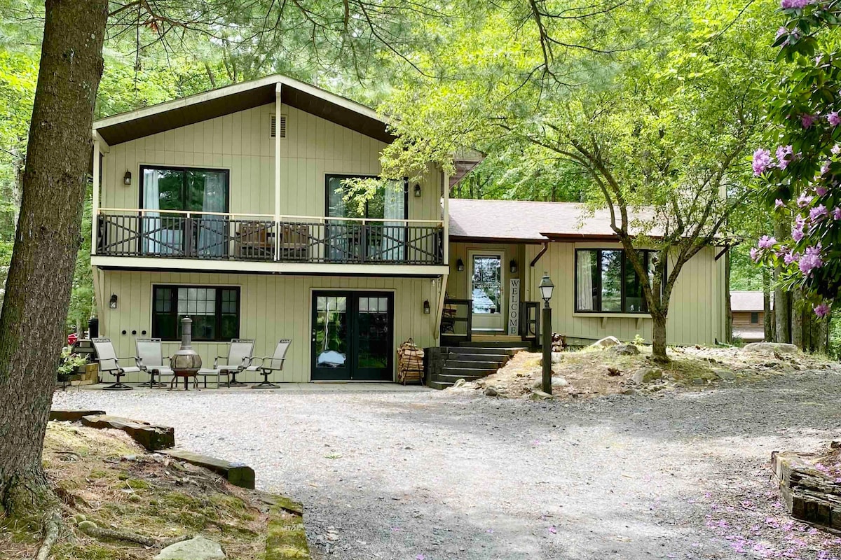 The exterior of the home showcases a two-story structure set among trees, featuring large windows on both floors. A front patio area includes seating for relaxation, while the pathway leads to the entrance. Surrounding landscaping features natural stone elements and greenery.