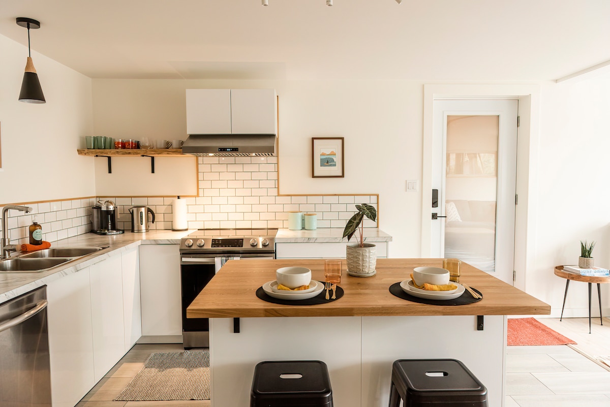A modern kitchen is presented with white cabinetry and stainless steel appliances. A wooden island with two stools invites casual dining. Natural light brightens the space, highlighting the tiled backsplash and countertop decor. A door leads to a bedroom area, subtly visible in the background.
