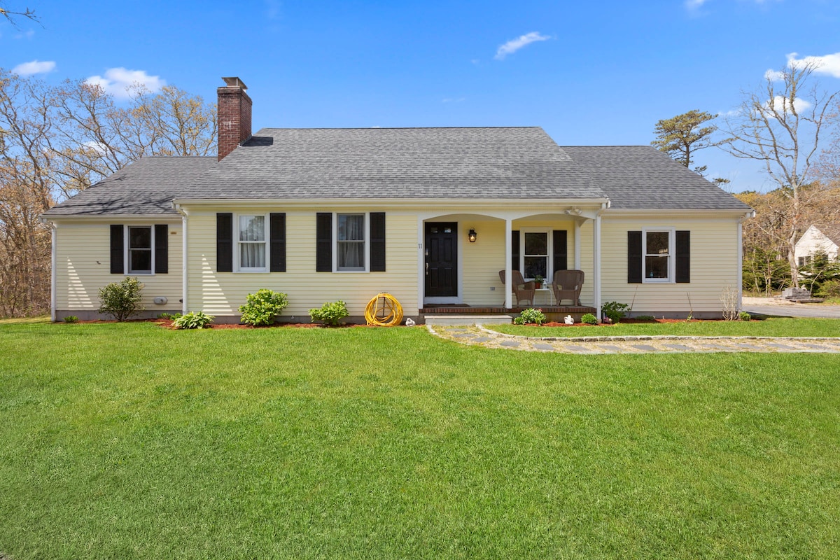 A charming Cape-style home is presented with a well-maintained front yard featuring lush green grass and neatly arranged flower beds. A stone pathway leads to the front door, flanked by two chairs and a welcoming door with a round window. Bright blue skies complete the scene.