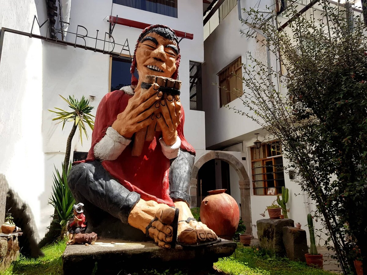 A large, colorful sculpture of a seated figure is displayed in an outdoor area, surrounded by greenery. The figure is depicted holding a small object, with details on the clothing and facial features visible. Light filtering through nearby buildings enhances the scene.
