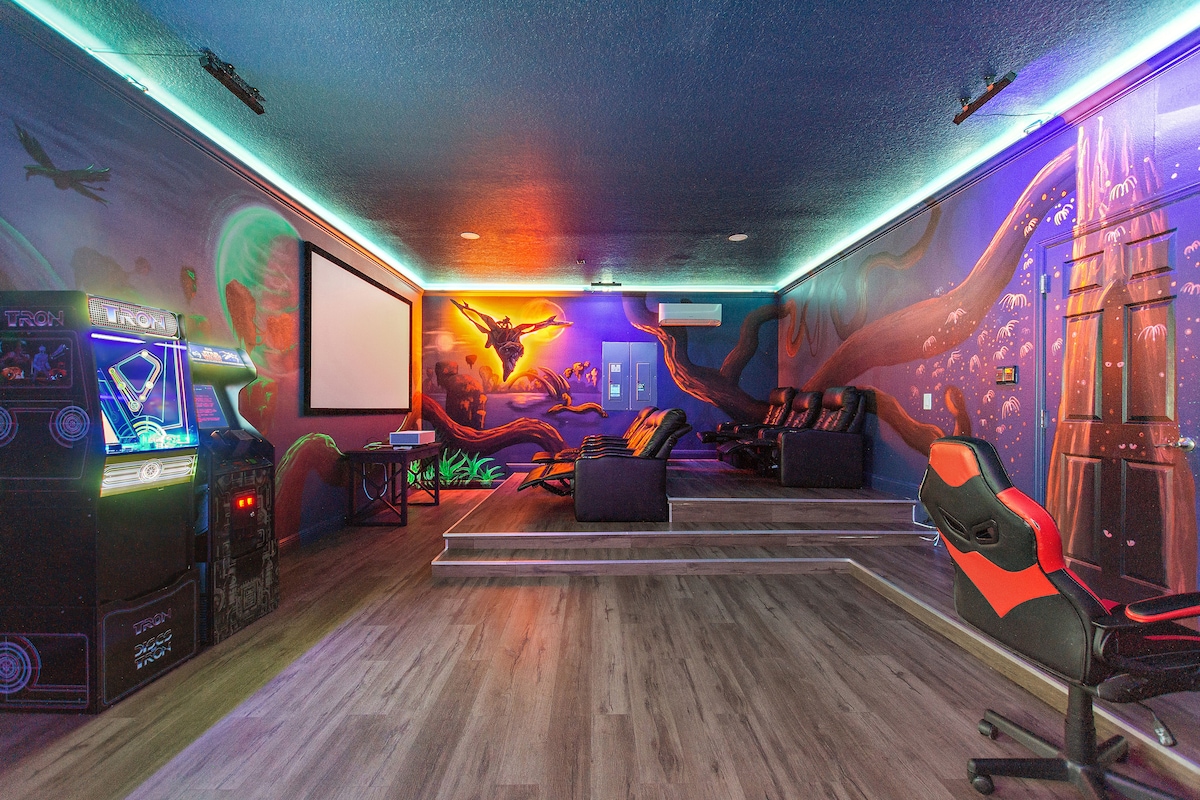 A vibrant game room features colorful murals covering the walls, depicting underwater scenes and fantasy elements. Comfortable seating is arranged in tiered levels for optimal viewing. An arcade game machine stands beside a projector screen, contributing to a fun atmosphere for guests of all ages.