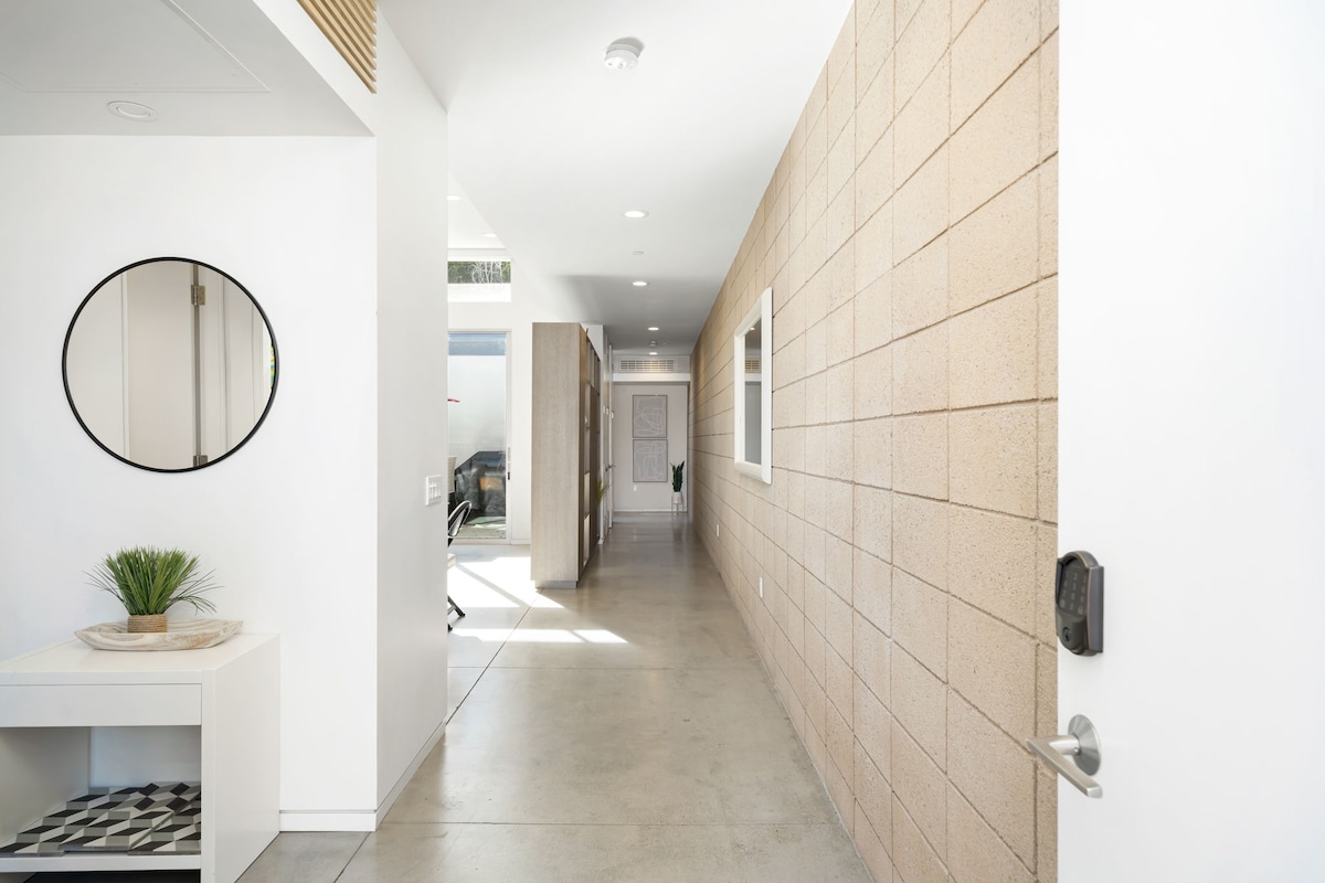 A spacious hallway is lined with smooth concrete flooring and light-colored walls. A round mirror is mounted on the left wall above a small console table with a decorative plant. Natural light is welcomed through windows, creating an open feel throughout the area.