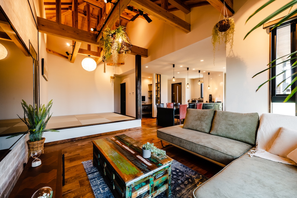 A spacious living area features high wooden beams and a mix of contemporary and rustic furnishings. An inviting sofa is paired with an artistic coffee table. Nearby, tatami space offers additional seating, and the background reveals an open dining area, enhancing the home's inviting layout.