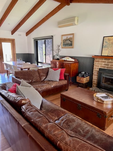 A comfortable lounge area features two leather sofas with accent pillows, facing a brick fireplace. Natural light enters through a large window, illuminating the warm tones of the wooden floor. A dining table is visible in the background, complementing the rustic décor.