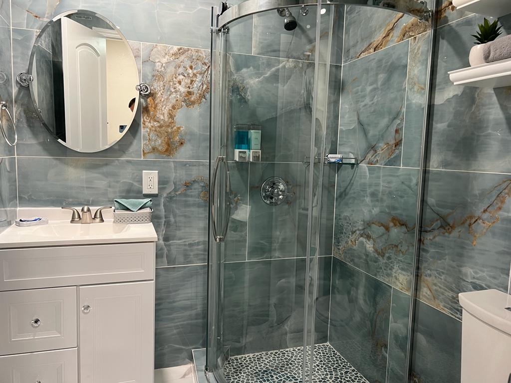 A modern bathroom features a glass shower stall with elegant marble-like tiles in shades of blue and gold. A round mirror is mounted above a white vanity, providing a bright and clean appearance. A small shelf with plants adds a touch of greenery.