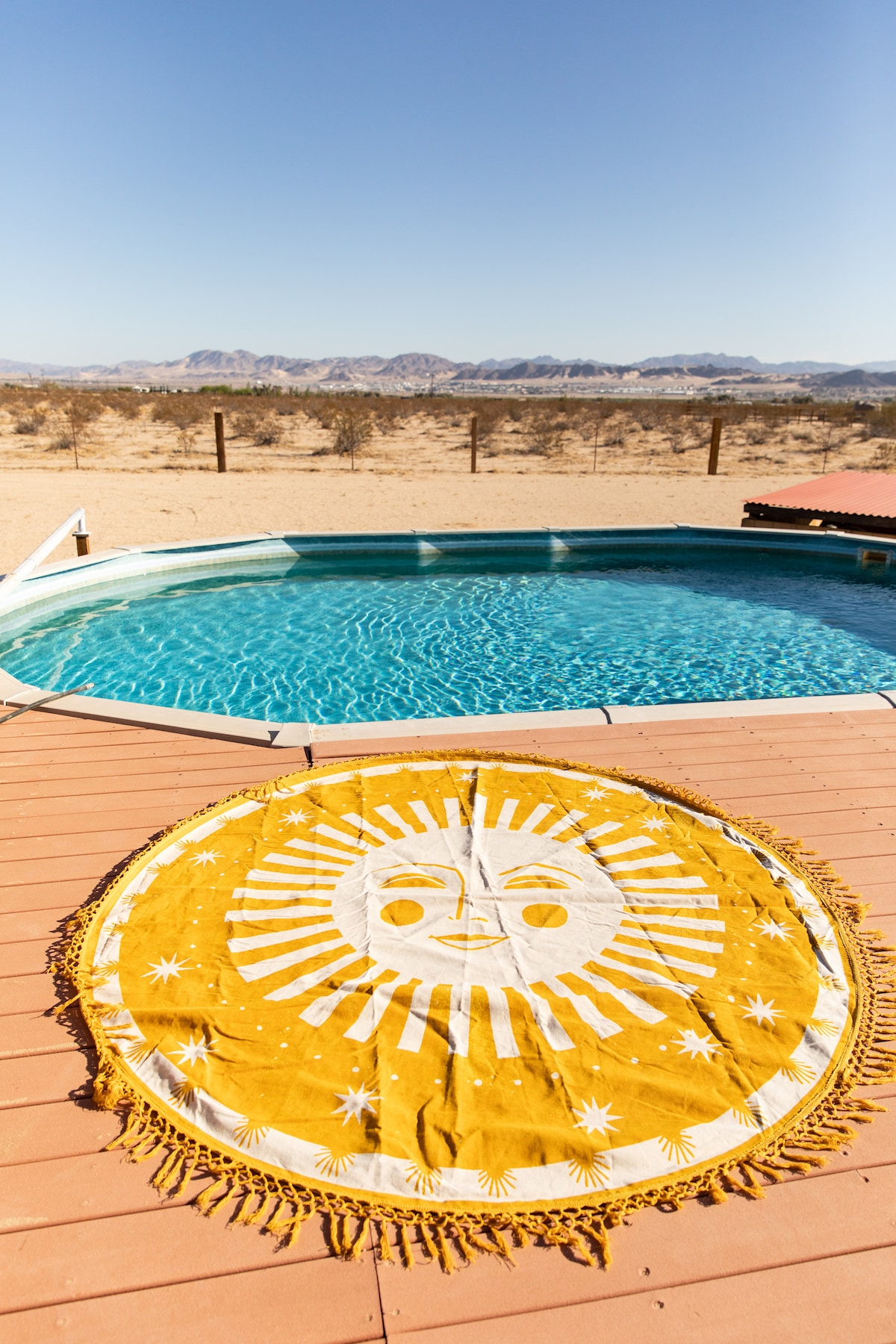 A circular sun-patterned blanket is spread on the poolside deck, complementing the inviting turquoise waters of the pool. The serene desert landscape stretches in the background, showcasing distant mountains under clear blue skies.