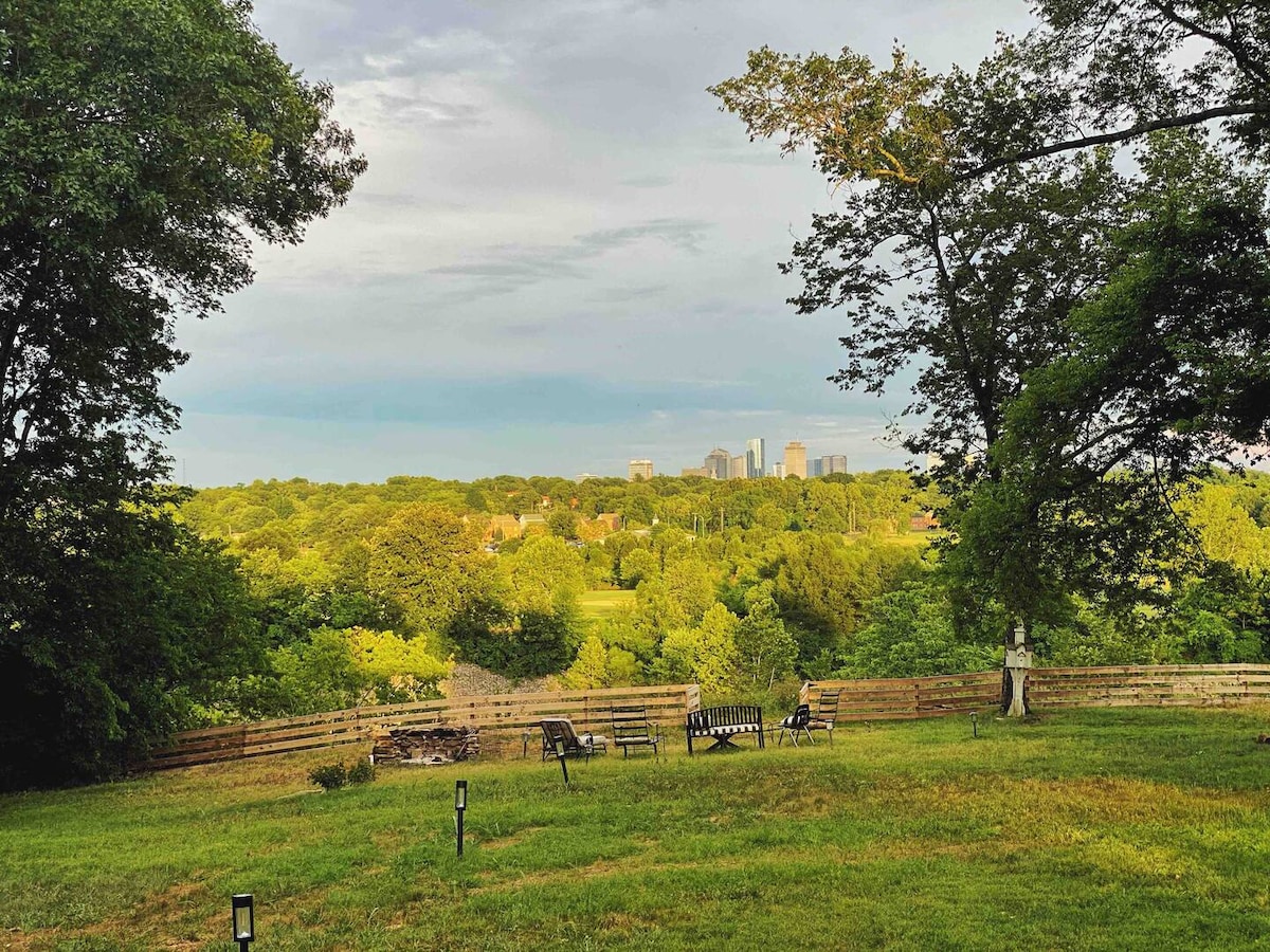 A spacious backyard is showcased, featuring green grass and a view of the Nashville skyline in the distance. Several outdoor chairs are positioned around a fire pit, providing a cozy gathering space beneath trees that offer shade and enhance the natural setting.