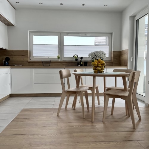 A bright kitchen area features modern cabinetry and a round glass dining table surrounded by four light-colored wooden chairs. Natural light floods through large windows, highlighting a centerpiece of lemons and flowers on the table.