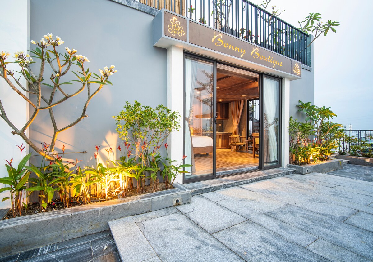 A modern exterior is framed by lush greenery and decorative plants, leading to spacious outdoor tiles. Large glass doors provide access to the interior, where warm wooden tones create a welcoming atmosphere. Soft lighting highlights the entrance, contributing to the overall inviting appearance.
