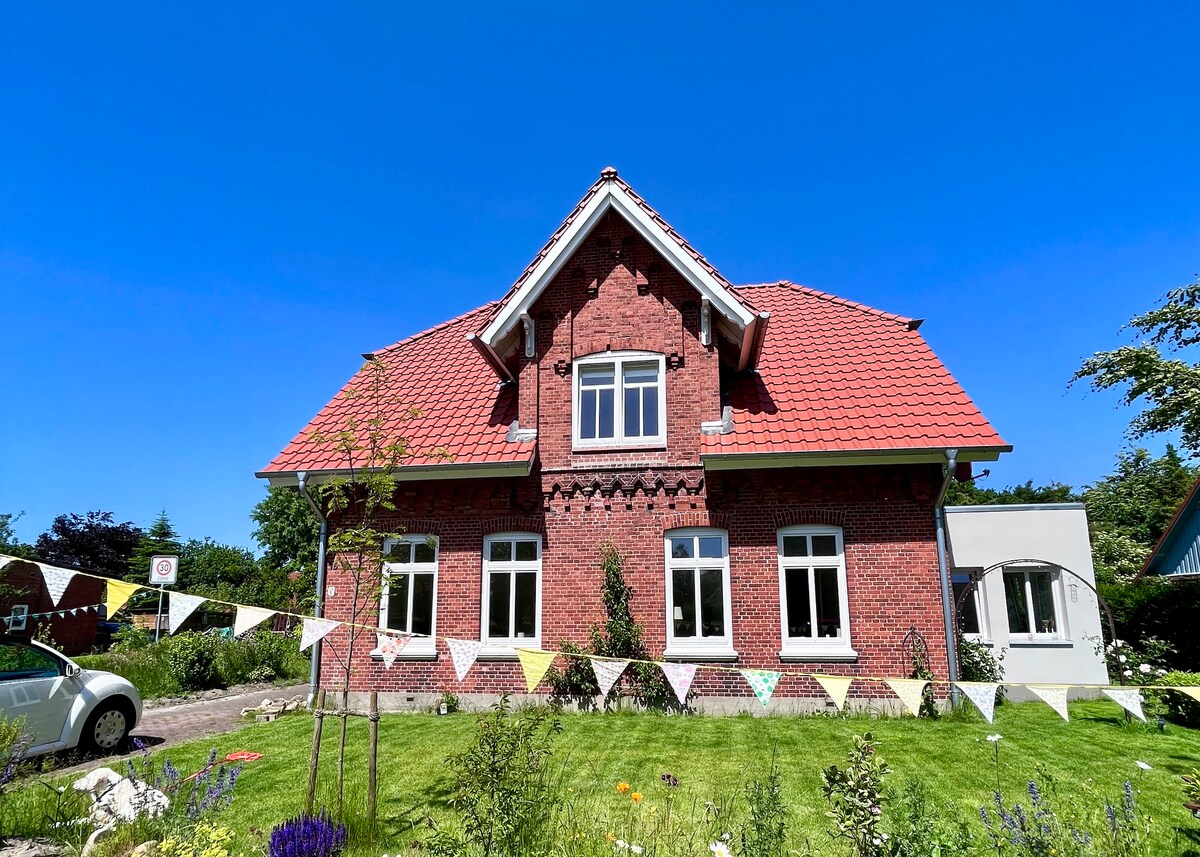 A charming red-brick house displays a classic roof design, set against a clear blue sky. Colorful bunting adds a festive touch along the garden area, which features green grass and blooming plants. Multiple windows allow natural light to brighten the interior.
