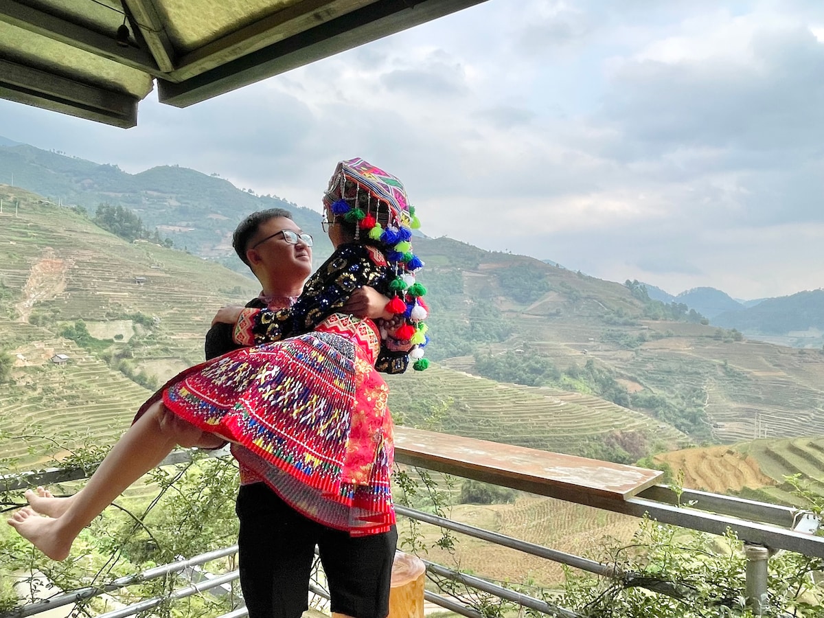 A couple is standing on a balcony overlooking terraced fields and rolling hills. The person being lifted wears a vibrant traditional outfit adorned with colorful pom-poms, while the landscape showcases varying shades of green under a cloudy sky.