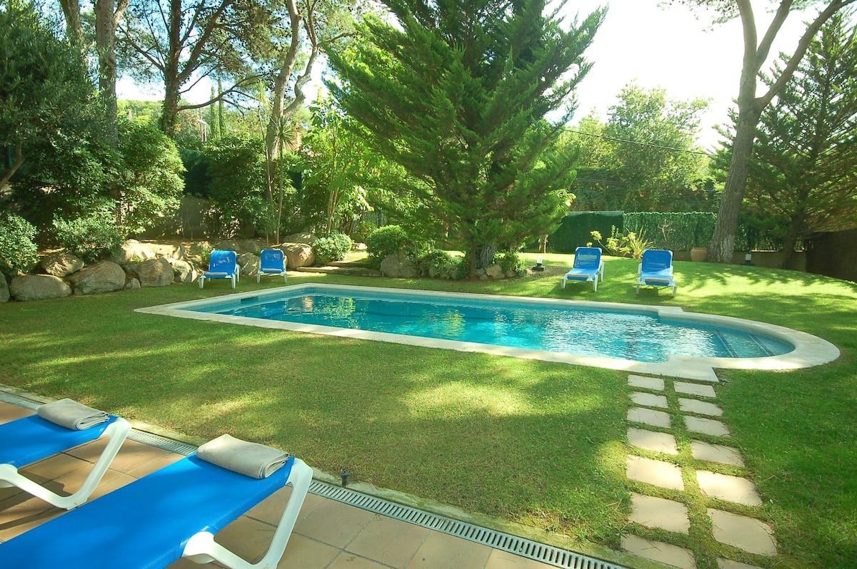 A serene garden area features a private pool surrounded by well-maintained grass. Blue sun lounges are positioned on one side, and a stone pathway leads to the pool. Tall trees provide shade, completing a tranquil outdoor atmosphere.
