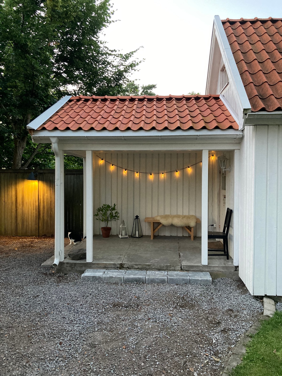 A covered patio features a light string illuminating the area. A small potted plant and a lantern are placed beside a bench with a soft throw. A black chair is positioned near the entrance, which is framed by white walls and a classic red-tiled roof.