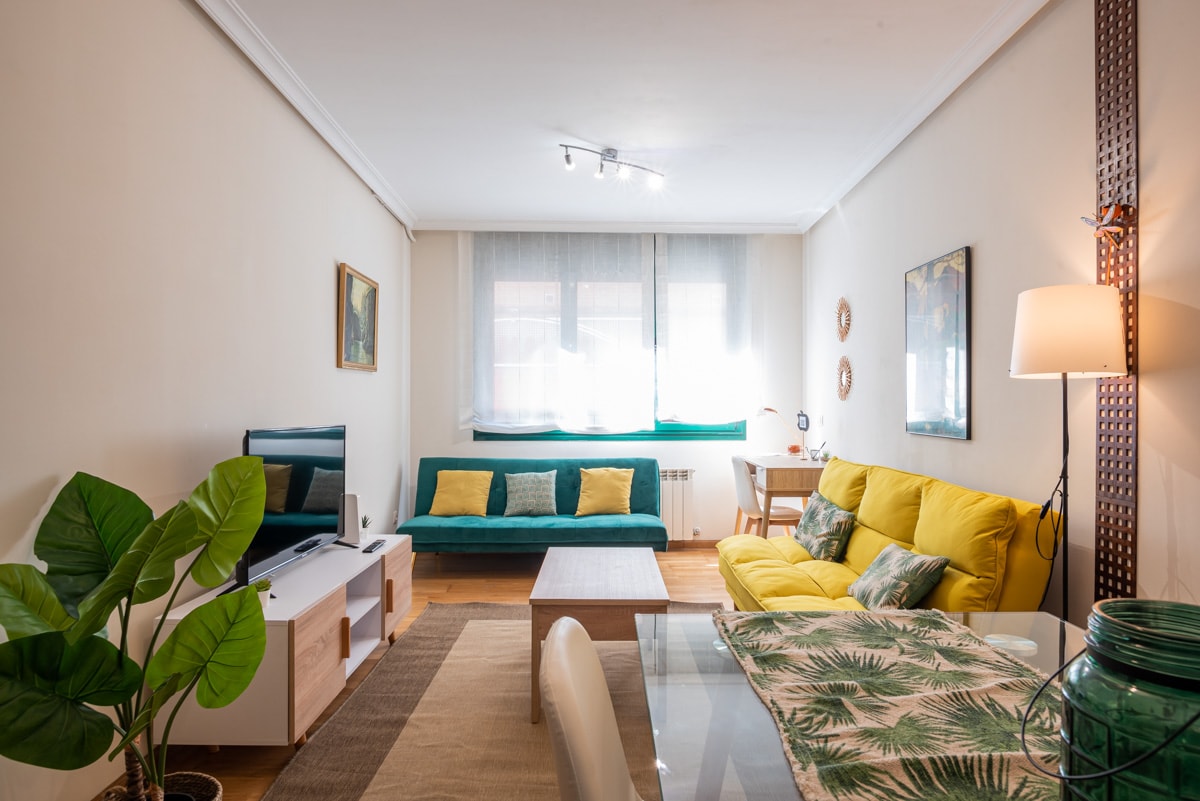 A bright living area is filled with natural light from large windows. A yellow sofa and a green sofa provide ample seating, complemented by a glass dining table. A decorative plant adds a touch of greenery, and wall art enhances the inviting ambiance.