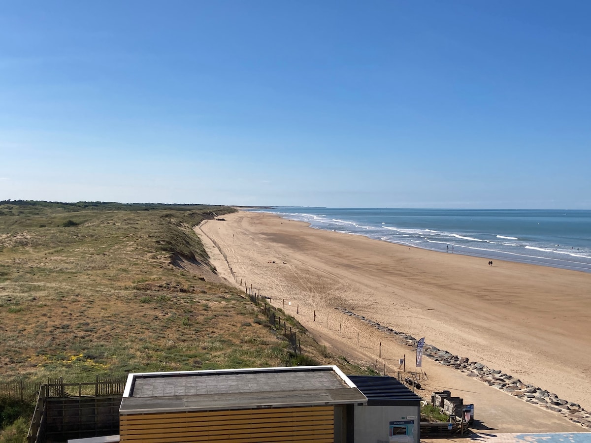 A wide sandy beach extends along the coastline, bordered by gentle dunes. The clear blue water meets the shore, and a few distant figures can be seen enjoying the beach. The sky is bright and clear, enhancing the serene coastal landscape.