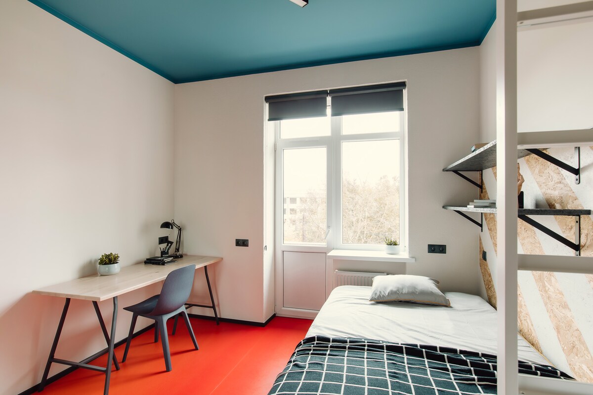 A well-lit room features a spacious bed with black and white bedding, complemented by a desk and chair for studying. Natural light enters through a large window, and a coffee maker sits on the desk. Blue and white accents add a modern touch.