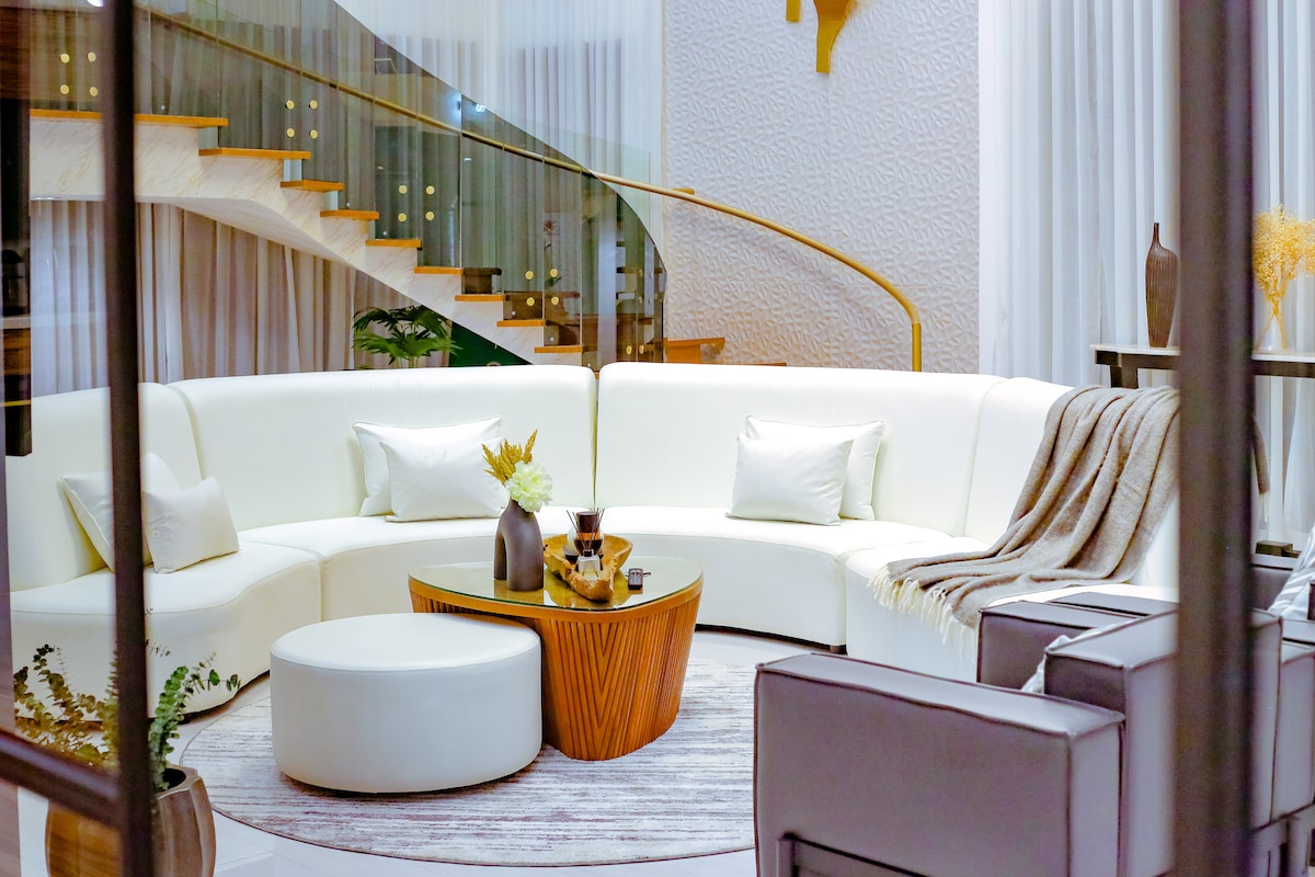 A spacious living area showcases a circular white sofa with soft cushions, complemented by a textured round coffee table. A plush throw is draped casually over the sofa. Warm lighting illuminates the space, featuring a stylish staircase visible in the background.