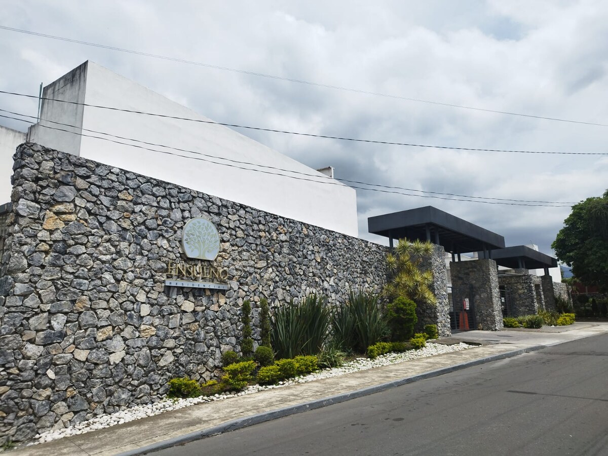 A stylish exterior view of a modern building framed by a natural stone wall. Lush greenery and decorative plants complement the entrance, which features a sleek canopy. The cloudy sky adds a muted backdrop to this welcoming structure.