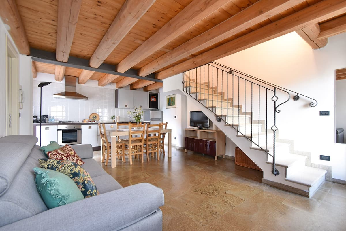 A spacious living area with wooden beams overhead enhances the contemporary style. An open kitchen with essential appliances is visible alongside a dining table, set for several guests. The comfortable sofa, adorned with colorful cushions, is positioned across from a television mounted on the wall.