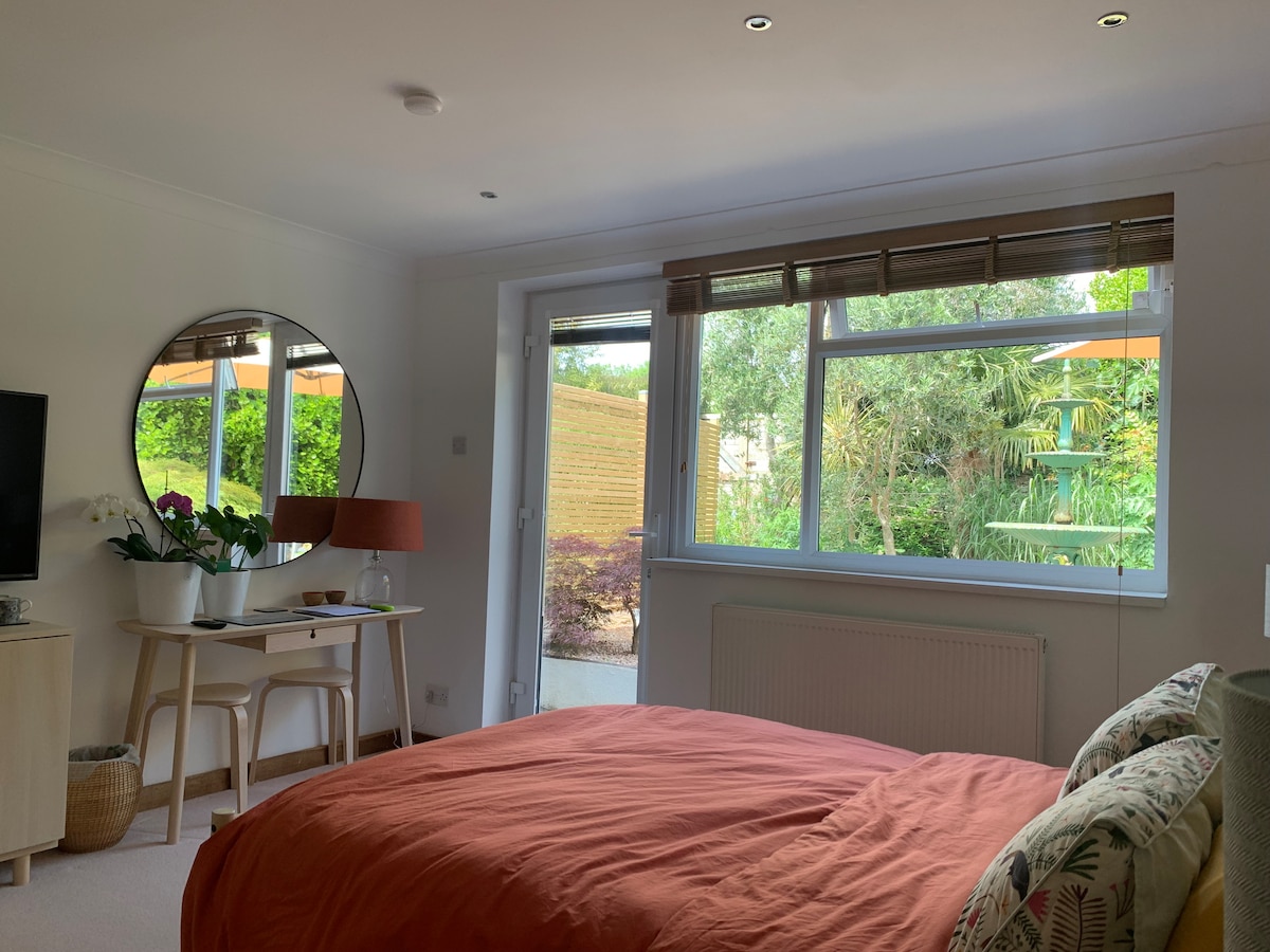 A stylish room is presented with a spacious bed covered in a soft orange duvet. A mirror reflects natural light from the large window, which offers views of the private garden. A small table with a lamp and a television are also visible.