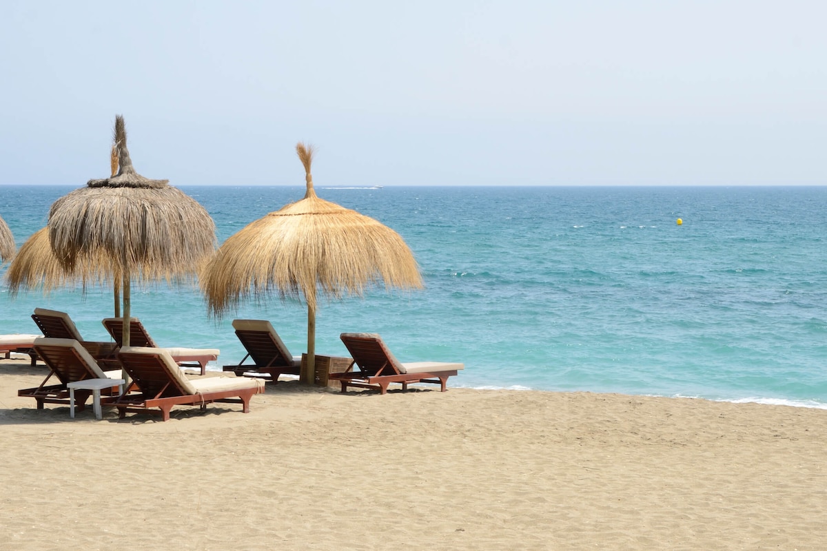 A tranquil beach scene features sun loungers arranged beneath thatched umbrellas. Soft golden sand extends towards a clear, turquoise sea. The horizon meets a light blue sky, creating a peaceful coastal atmosphere.