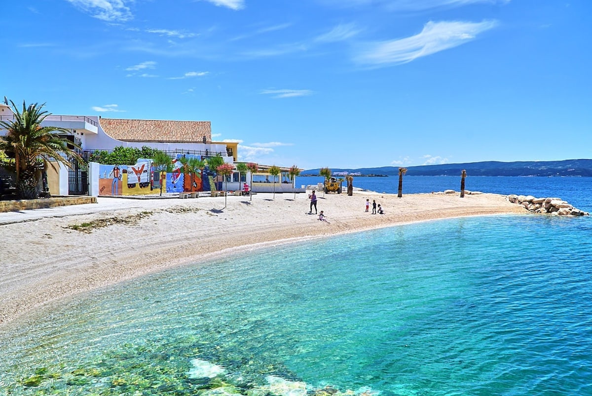 A sandy beach extends along the shoreline, bordered by palm trees and a charming building with murals. Clear blue waters reveal pebbles and rocks beneath the surface, while visitors stroll the beach, suggesting a tranquil atmosphere for leisure activities.